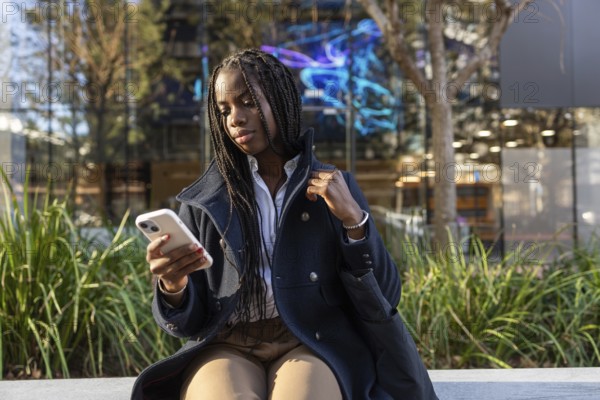 An African American businesswoman with braids intently uses a smartphone while seated outdoors on a work break, dressed in professional attire
