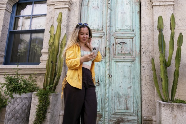 A female tourist stands in front of an ornate wooden door, intently using her smartphone to navigate or capture the moment. She wears a yellow jacket and sunglasses