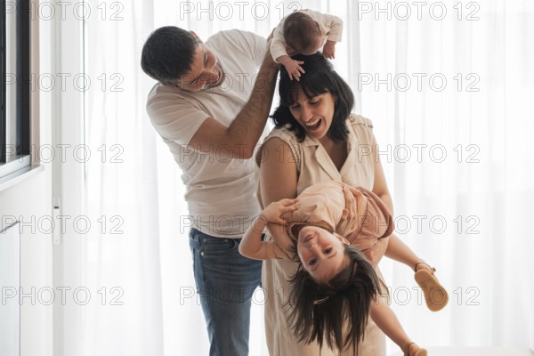 Happy young family shares joyful moments at home. The father holds a newborn while the mother playfully lifts their daughter. The big sister smiles upside down