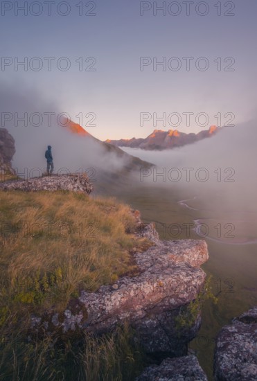 A lone hiker stands on a cliff, gazing at a stunning, mist covered valley at sunrise in the Pyrenees. The early light bathes distant peaks in golden hues, creating a tranquil scene in summer