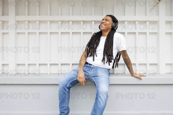 A joyful man with long dreadlocks and tattoos relaxes against a white fence, wearing headphones and a white shirt in Barranco, Lima, Peru, enjoying music