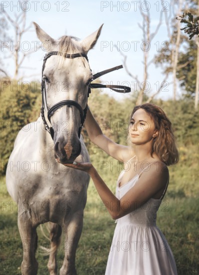 A young woman in a white dress gently touches a white horse face amidst a tranquil countryside background, showcasing the bond between human and nature