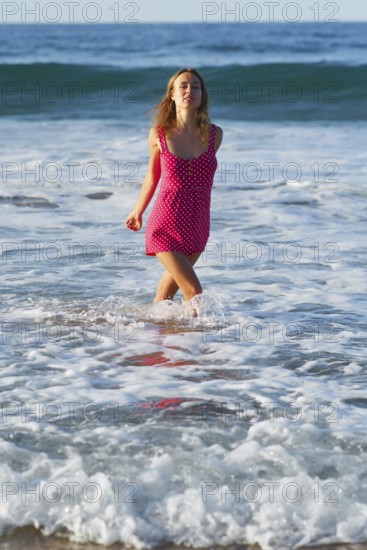 A woman in a vibrant pink polka dot dress joyfully runs through the surf at a sunlit beach in Zarautz, Spain, capturing the essence of a perfect summer day