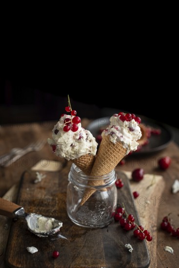 Two ice cream cones filled with creamy dessert, garnished with fresh currants, artistically placed in a glass jar on a rustic wooden table, accompanied by loose berries and a spoon