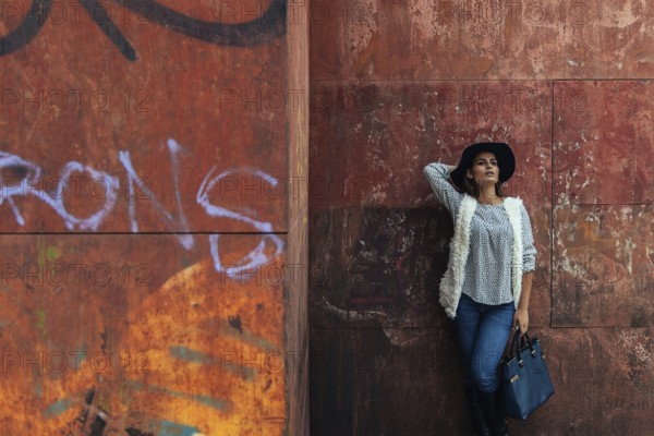 A stylish woman in casual attire with a hat leans against a graffiti-covered wall, showcasing urban fashion. The orange and red background adds a warm, textured contrast