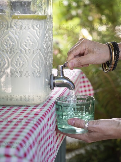 Fresh lemonade in large glass bottle with tap. Hands serving in glass. Refreshing summer drink. Liquid dispenser, sustainable and recyclable containers