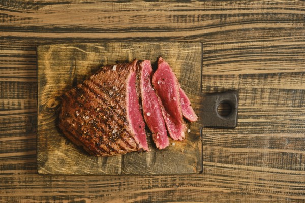Beef is sliced into thick pieces and placed on a wooden cutting board. The meat shows a reddish color and appears cooked. Seasoning is sprinkled on top. The background is a rustic wooden table