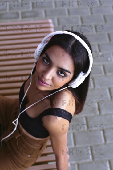 A young woman wearing white headphones sits on a wooden bench, looking up with a serene expression. The image captures a mix of relaxation and modern lifestyle outdoors