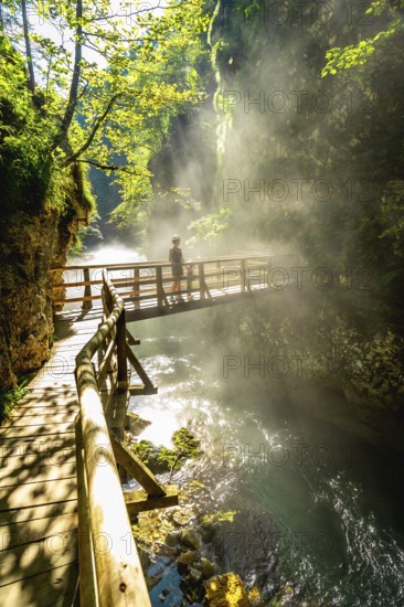 Sunbeams illuminating mist rising from radovna river in vintgar gorge near bled, with tourist walking on wooden footbridge