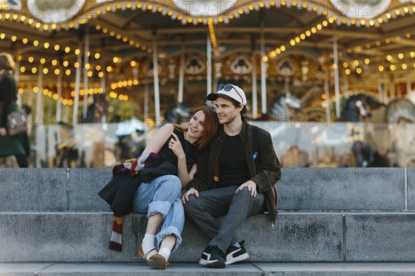 A couple sits on steps, embracing in front of a lit up carousel in Brooklyn Bridge Park. The joyful and warm scene captures a moment of togetherness