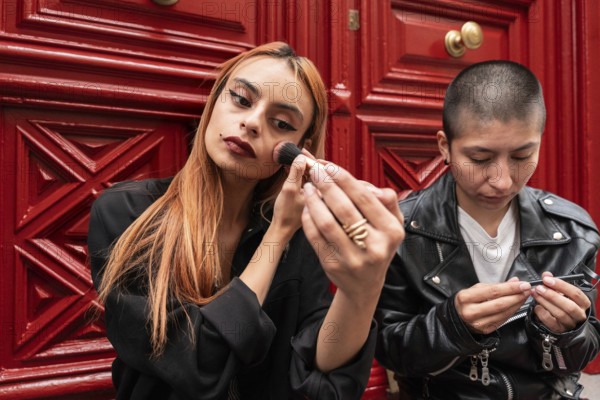 A lesbian couple enjoys a moment together, one applying makeup while the other engages in self-care, against a vibrant red backdrop, symbolizing love and individuality