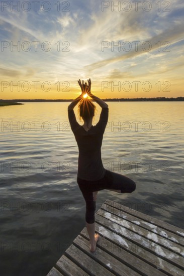 Woman practicing standing posture Vrikshasana, Tree Pose, one legged balancing asana of the medieval hatha yoga on jetty at lake at sunset
