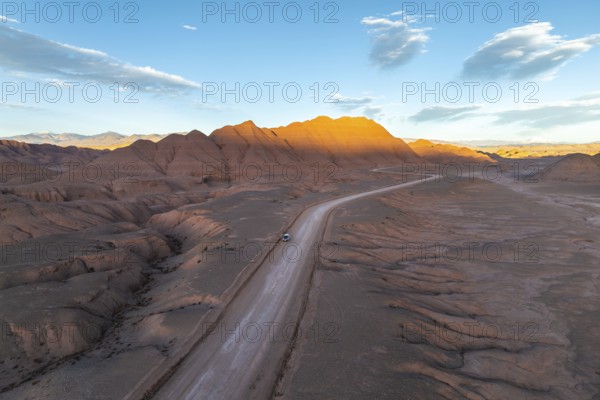 Aerial view of a vehicle in a empty desert road winding through rugged, sunlit mountains at dawn in the argentine Puna. The serene landscape offers a dramatic and peaceful panorama of natural beauty