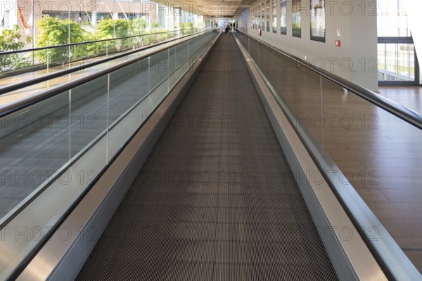 A modern airport travelator extends into the distance, surrounded by glass walls and natural light. The moving walkway provides convenience for travelers, enhancing airport transit