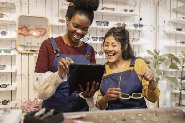 Two cheerful employees in a bustling glasses shop collaborate on a digital tablet, surrounded by a stylish array of eyewear, showcasing teamwork and customer service excellence