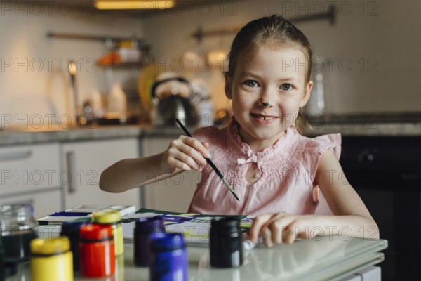A joyful child with a painted nose engages in a creative painting activity at home. She holds a brush while surrounded by colorful paint jars in a cozy kitchen setting