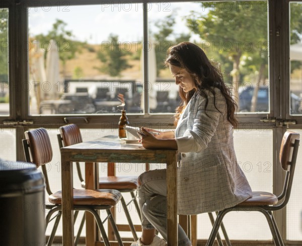 A woman sits in a sunny cafe, engaged with her tablet. Sunlight pours through the window, creating a warm ambiance. Relaxing and modern lifestyle scene