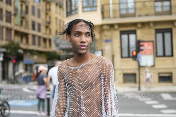 A confident man wearing a mesh top stands on a city street, embodying pride and LGBTQIA+ identity. Urban architecture forms an engaging backdrop