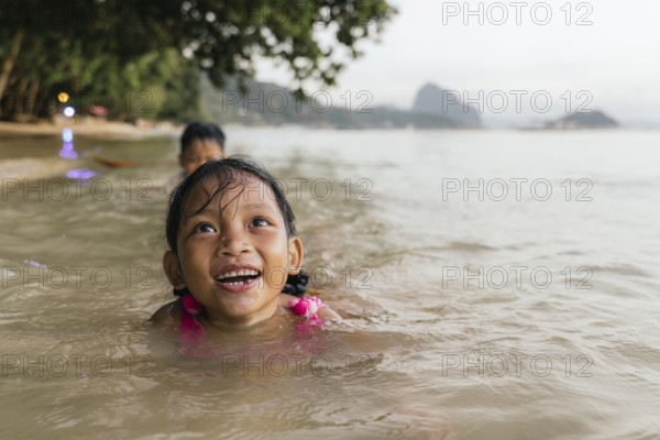 Two children enjoy swimming in the warm, shallow waters of a beach in the Philippines, surrounded by lush green trees, under a clear, bright sky, capturing the joy of carefree playtime