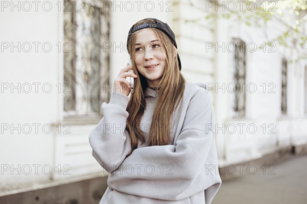 A teenage girl wearing a gray hoodie and black cap is happily speaking on her smartphone outdoors. She stands near a building, appearing relaxed and engaged in conversation