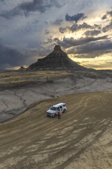 From above an unrecognizable person standing next to a SUV at Factory Butte, Utah, captured during a dramatic sunset with textured cloud-filled skies