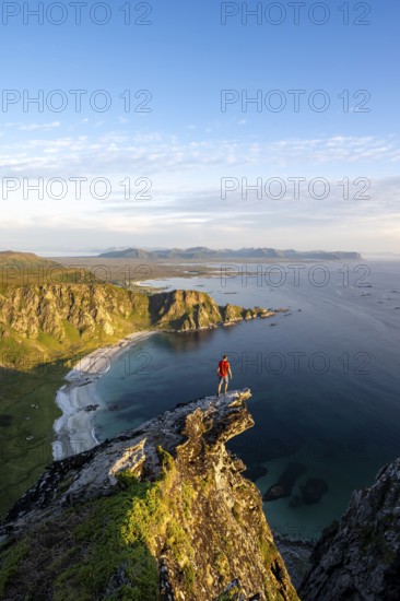 Evening mood, mountaineer standing on a rocky outcrop on the summit of Måtind, view of coastal landscape, Høyvika beach and sea, Måtinden, Andøya island, Vesterålen, Nordland Norway