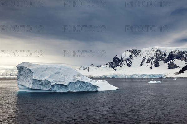An icy mountain floats in a wide, open sea in front of a dramatic mountain landscape, icebergs in the landscape of the Southern Ocean in Antarctica
