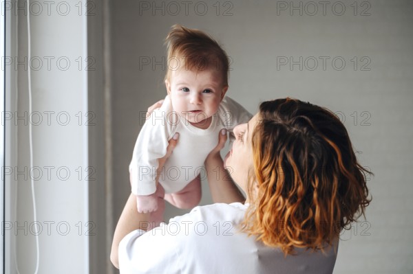 A mother lovingly holds her smiling newborn baby girl near a sunlit window, capturing a moment of joy and warmth. The little girl is dressed in a white onesie