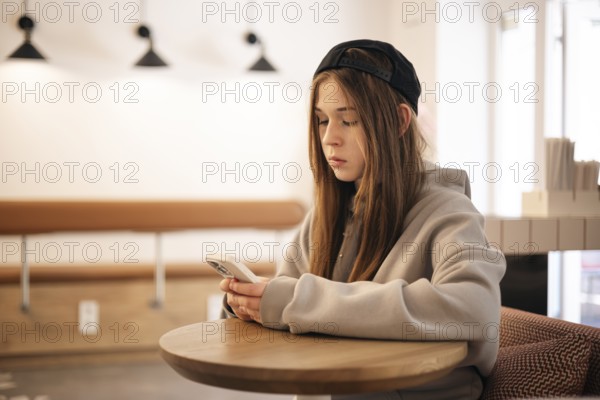 Teenage girl sitting at a cafe table using her smartphone. She is wearing a casual hoodie and cap, fully focused on the device in a modern, softly lit setting