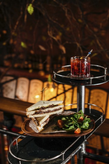 A delectable club sandwich with veal cheeks and cheese, paired with a refreshing Negroni cocktail. Accompanied by a fresh spinach and tomato salad on a wooden table