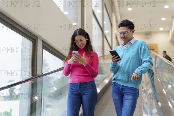 Young man and woman, Gen Z friends, enjoy using their smartphones while riding an escalator in a well-lit modern building, illustrating social media and digital connectivity