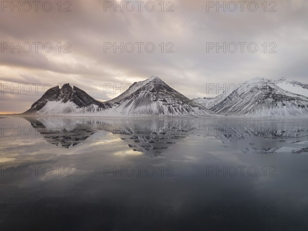Captivating view of Iceland's Stokksnes mountains reflecting on the calm, icy waters. Snow-dusted peaks meet the serene sky, creating a breathtaking winter scene
