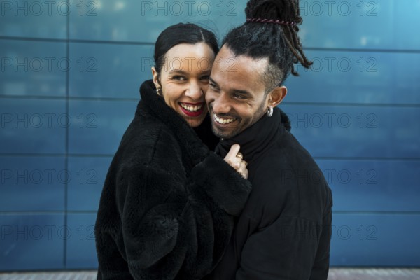 A multicultural couple stands close in a loving embrace, smiling warmly. The urban background adds depth to this candid portrait, highlighting love and diversity