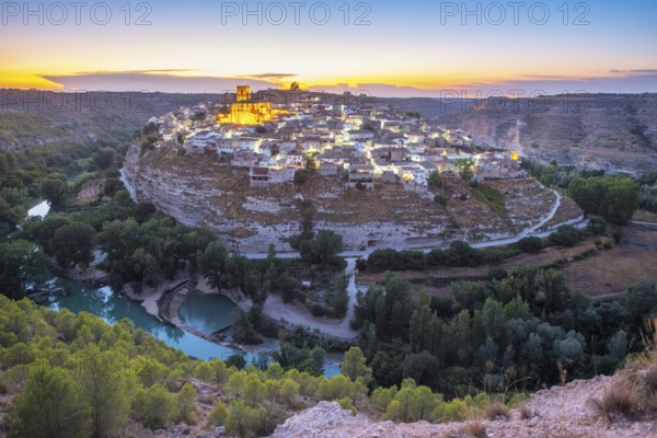 A breathtaking view of the historic town of Jorquera, Spain, perched on a cliff at sunset. The colorful houses and lush greenery are illuminated by the golden light of dusk