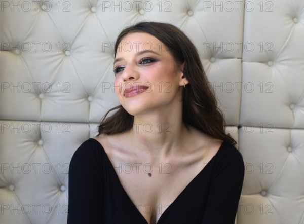 A woman with vibrant makeup and a subtle smile gazes thoughtfully. She is in an elegant setting with a padded wall backdrop, wearing a stylish black top