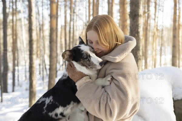 A redhead woman warmly embraces her dog, possibly a Border Collie mix, in a snowy forest setting. Both appear joyful and comfortable in their wintry surroundings