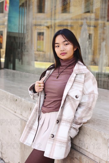 Young woman with a stylish plaid coat and pink blouse leaning against a stone ledge in an urban area, exuding confidence and modern fashion sense