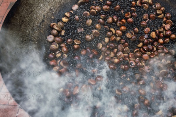 A traditional cooking scene from Hong Kong showing chestnuts being roasted in a large metal wok, with steam and smoke rising amidst the bustling atmosphere of a city market