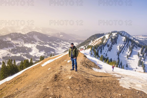 A hiker stands on a snow-covered mountain ridge with extensive mountain views under clear skies, Hochgrat Mountains, Allgäu Alps, Oberreute, Germany