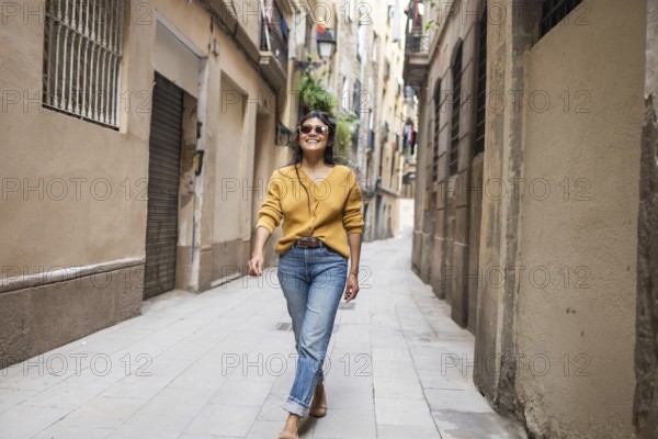 Latin woman in casual attire joyfully walks down a narrow, sunlit city street lined with old buildings. Her confident smile and relaxed demeanor create an inviting atmosphere