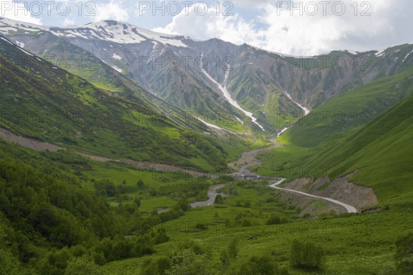 Green valley with snow-covered mountains under cloudy sky and a winding road, near Zagari Pass, Racha-Lechkhumi region and Lower Svaneti, Greater Caucasus, High Caucasus, Georgia