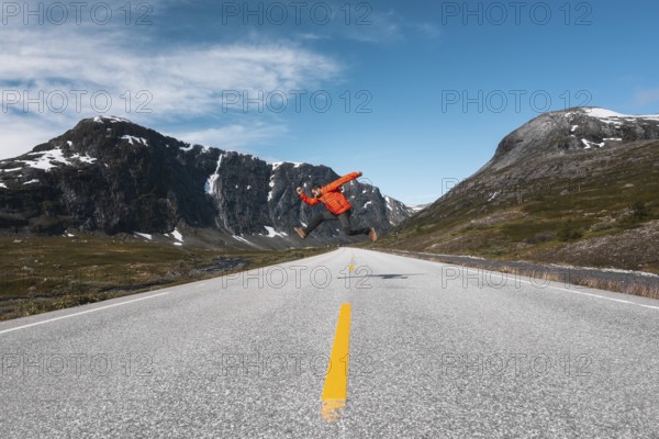 A traveler in a red jacket leaps mid-air on an open mountain road in Norway Framed by dramatic snow-capped peaks and a vibrant blue sky, this scene radiates adventure and freedom