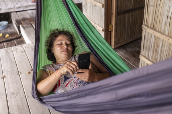 A mature woman enjoys a tranquil moment in a vibrant green hammock on Kri Island, Raja Ampat, while using her smartphone. This scene captures the essence of modern leisure in a rustic setting