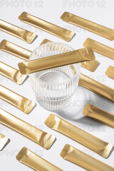 From above an esthetic arrangement of golden jelly collagen sachets with a clear glass resting on a smooth white background, capturing a modern and minimalist style