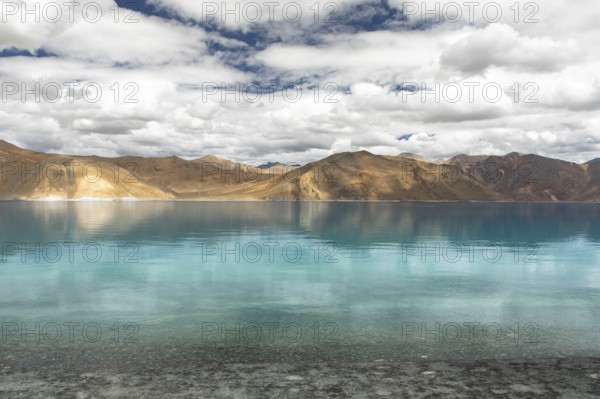A stunning view of Pangong Tso lake in Ladakh, India, with its crystal-clear turquoise waters reflecting the barren, rugged mountains under a partly cloudy sky