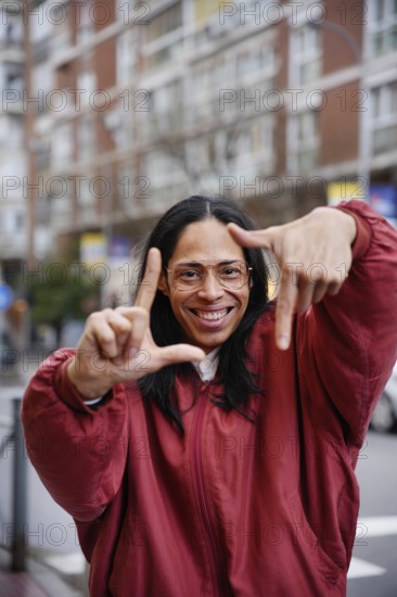 A joyful man in a red jacket creates a frame with their hands, emphasizing focus and perspective. The blurred urban background enhances the lively, bustling city ambiance