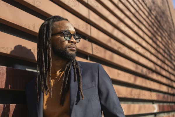 A cuban business man with sunglasses leans against a modern wall in sunlight, showcasing a blend of style and professionalism in an urban setting