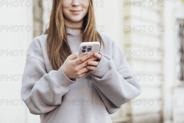 Teenage girl holding a smartphone while outdoors, dressed in a casual gray sweatshirt. The focus is on technology use and modern communication in everyday life