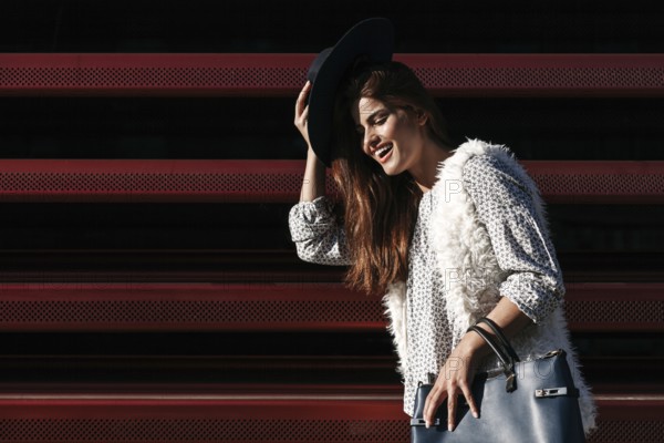 A fashionable woman poses outdoors on a sunny day, wearing a black hat, patterned shirt, and fur vest. She is holding a stylish handbag and smiling joyfully