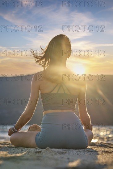 A woman sits in Padmasana on the beach, meditating with Chin mudra as the sun sets, creating a serene and calming atmosphere
